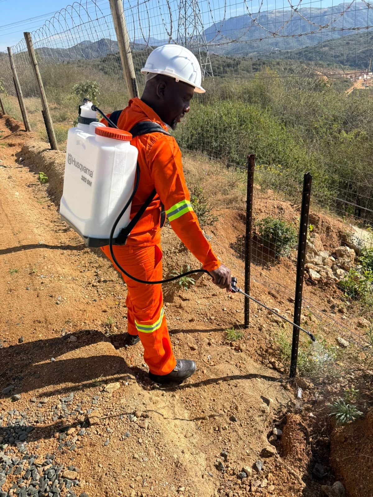 Shobelihle team member in full PPE carrying out vegetation control along a site perimeter fence